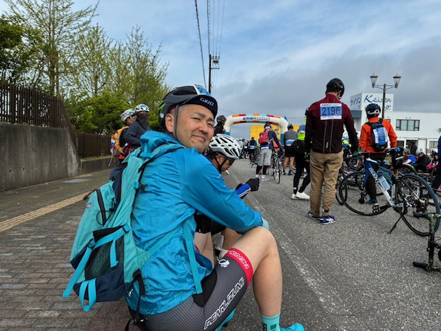Cyclist in blue jacket and helmet sitting on curb with backpack during a cycling event, others with bikes and an inflatable arch in the background