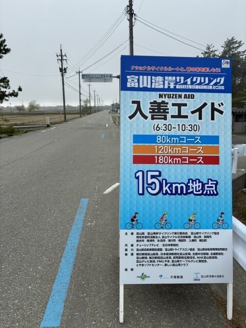 Blue roadside sign advertising a cycling event (Nyūzen Aid) with 15 km checkpoint and 80/120/180 km routes; cyclists pictured at the bottom on a rural road.