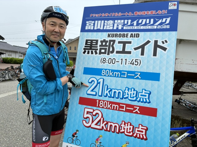 Cyclist in a blue jacket and helmet standing beside a large blue event sign, giving a thumbs-up; sign shows Toyama Bay Cycling event details with 80km course, 21km point, 180km course, and 52km point.