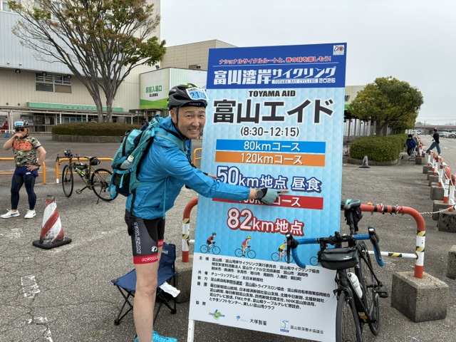 Cyclist in blue jacket and helmet posing beside a large event sign with 80km and 120km cycling routes at a Toyama aid station
