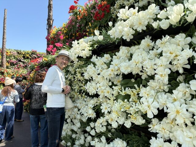 A man in a light sweater and hat stands beside a wall of white blooming flowers, with visitors in the background at a garden or flower market.