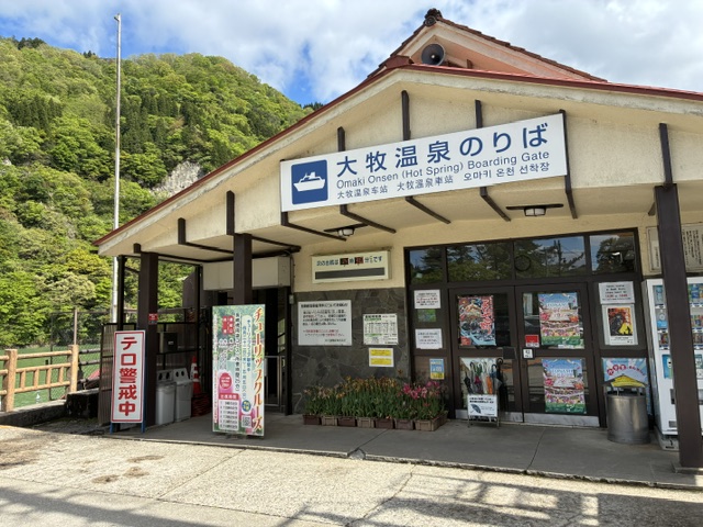 大牧温泉遊覧船乗り場 | 会社と社員と自分も成長させるのが、私の仕事 Building with a blue and white Omaki Onsen boarding gate sign next to glass doors, vending boards outside, surrounded by green hills.