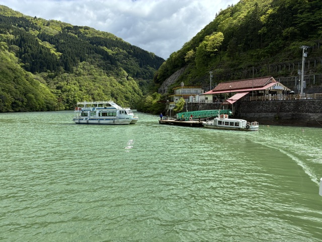大牧温泉遊覧船 | 会社と社員と自分も成長させるのが、私の仕事 Green river with two white boats near a forested hillside and a red-roofed riverside building.