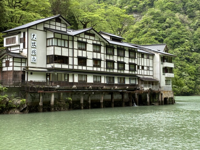 大牧温泉 | 会社と社員と自分も成長させるのが、私の仕事 Traditional Japanese ryokan (inn) perched over a green lake, with wooden stilts and a sign in Japanese characters on the facade.