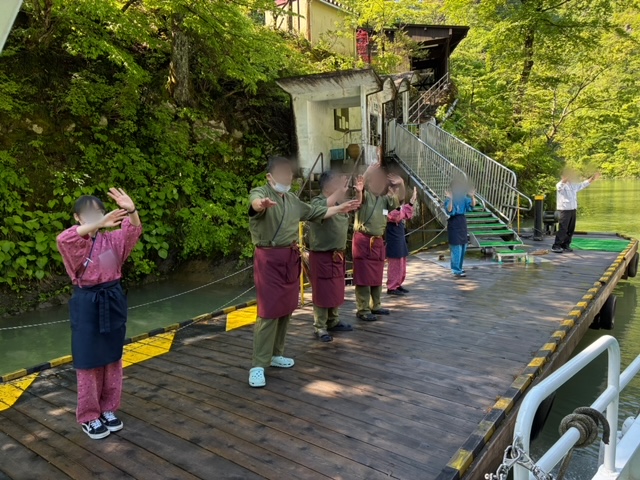 Group of people in matching aprons on a wooden dock by a green river, raising their hands.