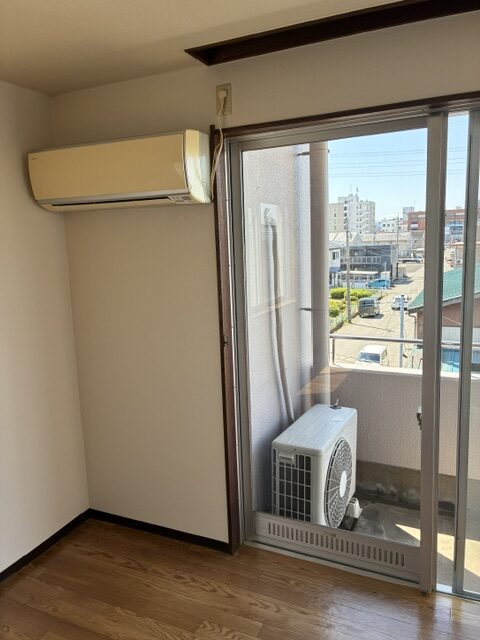 Empty corner room with a wall-mounted air conditioner and a sliding glass door to a balcony; outdoor unit resting on the balcony floor with a city street beyond.