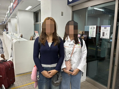 Two women with blurred faces stand in an airport baggage area next to luggage; one wears a navy sweater and jeans, the other a white jacket.